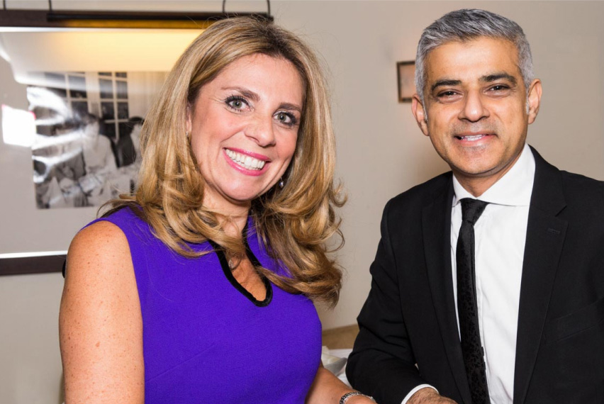 A woman, Nicola Mendelsohn, wearing a purple sleeveless dress, is shaking the hand of a man, Sadiq Khan, who wears a black jacket & tie with a white shirt. Both are smiling and looking directly to camera.